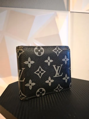 Svart monogram plånbok - Snygg svart plånbok med klassiskt monogrammönster i grått. Tillverkad i tåligt läder med flera fack för kort och sedlar. Perfekt accessoar för dig som gillar lyxiga detaljer och tidlös design.
