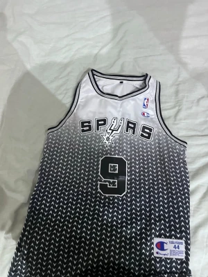 San Antonio Spurs basketlinne #9 - Snyggt basketlinne från Champion med San Antonio Spurs tryck och nummer 9, Parker på ryggen. Linnet är vitt upptill och övergår i svart med pilmönster. NBA-logga på bröstet och klassisk sportig design. Perfekt för basketfans!
