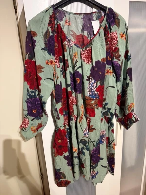 Blommig kortklänning med v-ringning - Snygg kortklänning med v-ringning och trekvartsärm. Klänningen har ett blommigt mönster i rött, lila och grönt på ljusgrön botten. Perfekt för dig som gillar färg och mönster. Materialet känns lätt och luftigt.