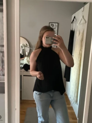 Svart halterneck topp - Säljer en svart halterneck topp med hög hals och ärmlös design. Toppens passform är loose och den är perfekt att matcha med jeans eller kjol. Materialet känns lätt och luftigt, vilket gör den bekväm att bära under varma dagar.