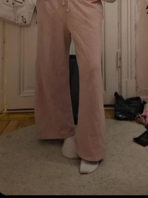 Ljusa rosa mjukisbyxor med vida ben - Mjukisbyxor i ljusrosa färg med breda, raka ben och dragsko i midjan. De är tillverkade i ett mjukt och bekvämt material som passar perfekt för chill dagar. Byxorna har en avslappnad passform och är riktigt sköna att ha på sig.