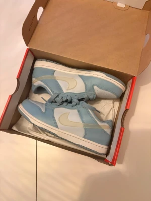Nike Dunk Low Aura/Blue-White 35 - Nike Dunk Low sneakers i färgen Aura/Clear-Worn Blue-White. Skorna har en klassisk låg siluett med ljusblå och vita paneler, vit sula och ikonisk Swoosh i vitt. Tillverkade i läder och textil med matchande blå skosnören. Perfekta för dig som gillar stilrena sneakers.