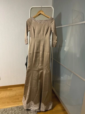 Beige långklänning med spetsdetaljer - Elegant beige långklänning med trekvartsärm och vacker spetsbroderi vid halsringning och nederkant. Klänningen har en klassisk siluett och är tillverkad i ett glansigt material som ger en lyxig känsla. Perfekt för dig som vill ha en stilren och sofistikerad look.