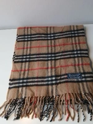 Beige rutig halsduk från Burberrys - Klassisk beige halsduk från Burberrys med ikoniskt rutmönster i svart, vitt och rött. Tillverkad i 100% lammull och har fransar i båda ändar. Perfekt accessoar för att lyfta din outfit med en tidlös brittisk touch.
