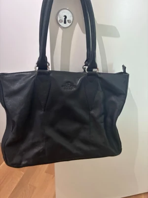 Svart läderhandväska från Tiger - Stilren svart handväska i äkta läder från Tiger. Väskan har dubbla långa handtag, silverfärgade metalldetaljer och diskret logga framtill. Rymlig modell med mjuk form som passar perfekt till alla dina essentials.