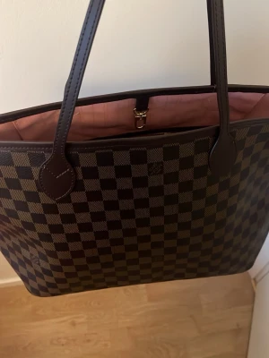 Brun väska - Snygg shoppingväska. Väskan har mörkbruna läderhandtag och rosa inner. Perfekt för dig som vill ha en lyxig och rymlig väska med ikonisk design. Aldrig använd. Bra material. Skriv om du har frågor.