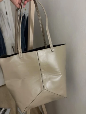 Beige shoppingväska från H&M - Snygg beige shoppingväska från H&M i slätt konstläder med dubbla axelband och rymligt fack. Väskan har en stilren design med dekorativa sömmar framtill och en innerficka för småsaker. Perfekt för att bära allt du behöver under dagen.