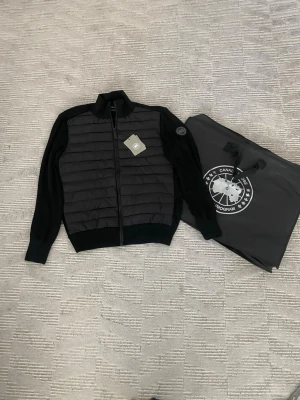 Svart canada goose cardigan st L - Snygg svart quiltad jacka från Canada Goose med ribbade muddar och dragkedja framtill. Jackan har långärmade stickade ärmar och en hög krage. Perfekt för kyliga dagar.