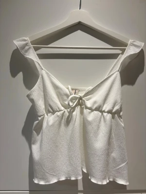 Vit linnetopp med knyt fram - Söt vit linnetopp med knyt fram och breda axelband. Toppen har en lätt strukturerad yta och luftig passform, perfekt för varma dagar. Passar dig som gillar en enkel men ändå stilren look. Mer off white än vit. 💖fri frakt till söndag 26  april