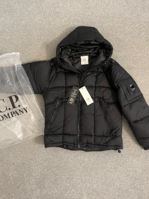 Svart pufferjacka från C.P. Company - Svart pufferjacka från C.P. Company med huva, två dragkedjeförsedda fickor och ikonisk linsdetalj på ärmen. Jackan är quiltad och tillverkad i ett glansigt syntetmaterial som håller värmen under kalla dagar. Perfekt för vinter och streetwear-stil.