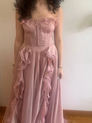 Lång rosa klänning med volanger - Romantisk långklänning i ljusrosa med volangdetaljer och knappar framtill. Axelbandslös design med markerad midja och svepande kjol. Perfekt för bal eller speciella tillfällen. Klänningen passar XS men också S.