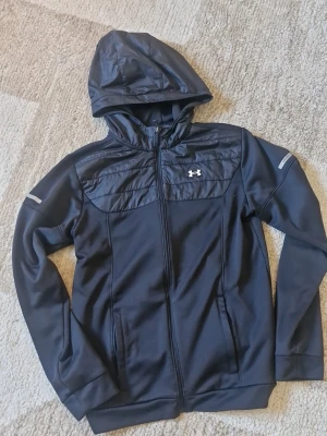 Svart hoodie från Under Armour - Cool svart hoodie från Under Armour med quiltad detalj över bröstet och huva. Materialet är en mix av syntet och fleece, perfekt för att hålla värmen. Dragkedja framtill och två sidofickor. Diskreta reflexdetaljer på ärmarna. Fodrat material innanför jackan och tål mycket blåst och vindar. Inte ett förhandlingsbart pris för en så pass oanvänd under armour jacka. Kom pv för fler bilder!