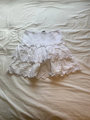 Vit volangkjol från BikBok - Supersöt vit kortkjol från BikBok med bred smock i midjan och två lager volanger med spetsdetaljer. Perfekt för dig som gillar en romantisk och feminin stil. Kjolen är gjord i bomull och har en luftig känsla. Har bra använt kjolen en gång och säljer den för att den inte kommer i användning. Köpte kjolen för 400kr