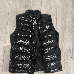 Svart dunväst från Moncler, S - Snygg svart dunväst från Moncler med glansig finish och hög krage. Västen har dragkedja framtill, två fickor med dragkedja och klassisk Moncler-logga på bröstet. Perfekt för lager-på-lager och streetstyle.