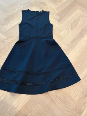 Mörkblå ärmlös klänning med spets - Snygg mörkblå klänning med ärmlös design och rund hals. Kjolen har fina spetsdetaljer och markerad midja som ger en feminin siluett. Perfekt för dig som gillar stilrena plagg med en twist. Materialet känns mjukt och följsamt.