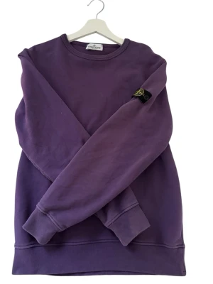 Lila sweatshirt från Stone Island - Säljer en lila sweatshirt från Stone Island med klassisk rund hals och ribbade muddar. Tröjan har den ikoniska Stone Island-loggan på vänster ärm och är gjord i mjukt bomullsmaterial. Perfekt för dig som gillar streetwear och vill ha en stilren look. Hör av er vid fler frågor eller funderingar 