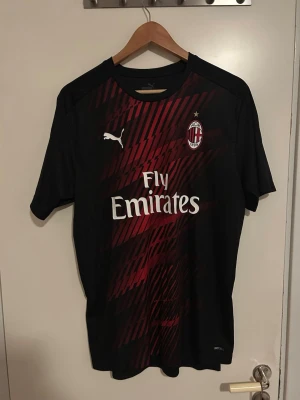 AC Milan matchtröja Puma svart/röd - AC Milan matchtröja från Puma i svart med röda diagonala mönster på framsidan. Tröjan har klubbmärke, Puma-logga och Fly Emirates-tryck på bröstet. Korta ärmar och sportig passform i lätt funktionsmaterial.