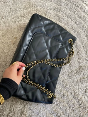 Svart quiltad axelväska  - Snygg svart axelväska i quiltat konstläder med guldfärgad kedjerem. Väskan har en mjuk, puffig look och klassisk design som passar till många outfits. Perfekt för dig som vill ha en trendig och stilren accessoar.