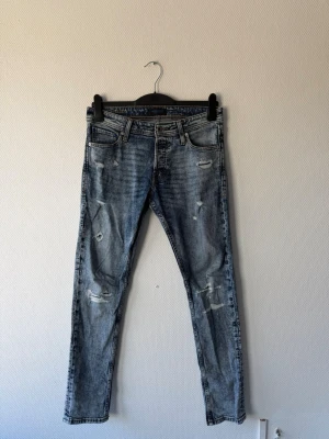 Jack & Jones jeans - Säljer ett par blå slim fit jack & jones jeans med slitningar och distressed detaljer på benen. Jeansen har klassisk femficksdesign och är tillverkade i denim med en snygg tvättad look. Perfekt för dig som gillar en trendig och avslappnad stil.