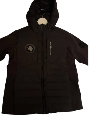 Svart dunjacka från Sail Racing - Svart dunjacka från Sail Racing med huva och dragkedja framtill. Jackan har två sidofickor, logotyp på bröstet och patch på ärmen. Lång modell med quiltade partier och hög krage för extra värme. Hör av er om ni har frågor.