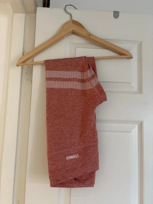 Rosa seamless leggings STRONGER - Säljer ett par rosa seamless leggings från STRONGER med vita ränder och logga på benet. De har hög midja och är tillverkade i ett stretchigt material som sitter skönt på kroppen. Perfekta för träning eller chill dagar.