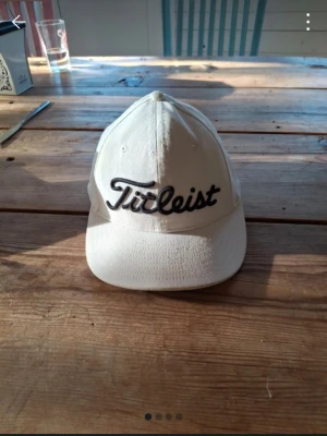Vit keps från Titleist - Snygg vit keps från Titleist med klassisk böjd skärm och broderad logga både fram och bak i svart. Kepsen är tillverkad i bomull och har ventilationshål för extra komfort. Perfekt för dig som gillar sportig stil och golf. Size S/M