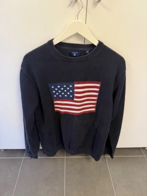 Mörkblå stickad tröja GANT USA-flagga XL - Mörkblå stickad tröja från GANT med stor amerikansk flagga broderad på bröstet. Rund halsringning och ribbade muddar. På ena ärmen står det 'GANT NH CT 1949' broderat i vitt. Perfekt för dig som gillar klassisk college-stil.
