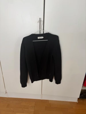 Svart cardigan från Weekday - En klassisk svart cardigan från casual friday med v-ringning och knappar framtill. Tröjan har långa ärmar och är gjord i ett mjukt stickat material som är perfekt att slänga på sig när det är lite kyligt. Enkel och stilren design som funkar till många outfits.