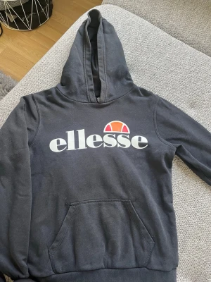Svart Ellesse hoodie med logga - Säljer en svart hoodie från Ellesse med stor vit och orange logga på bröstet. Tröjan har huva och en klassisk magficka. Perfekt för dig som gillar sportig och avslappnad stil.