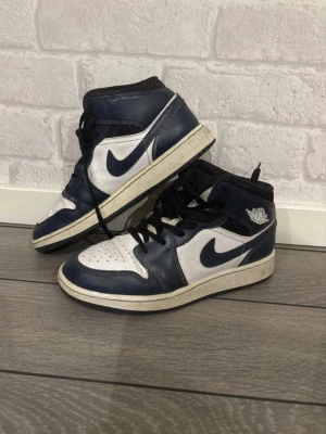 Nike Air Jordan 1 Mid blå/vit - Säljer ett par Nike Air Jordan 1 Mid sneakers i blått och vitt läder med svarta detaljer och klassisk Air Jordan-logga på sidan. Skorna har svart snörning och vit sula med blått swoosh-märke. Perfekta för dig som gillar streetwear och ikoniska sneakers.