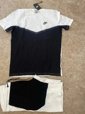 Nike svartvit body och shorts set - Säljer ett snyggt set från Nike med en svartvit body och matchande shorts. Bodyn har en cool V-formad färgblockdesign med svart nedre del och vit övre del samt Nike-logga på bröstet. Materialet är mjukt och bekvämt, perfekt för att chilla eller träna.