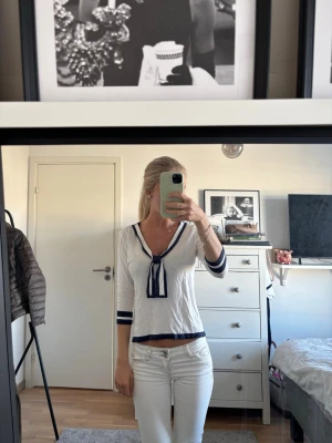 Vit blus med marinblå detaljer - Vit blus med marinblå kantband och rosett framtill. Blusen har v-ringad hals och trekvartsärmar med blåa ränder vid ärmslut och nederkant. Perfekt för en stilren och klassisk look.