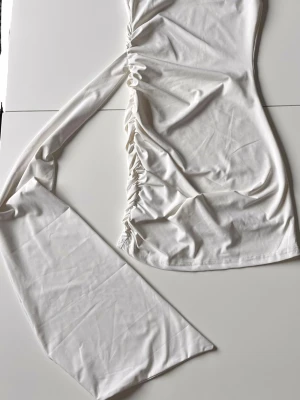 Vit kortklänning med snörning - Trendig vit kortklänning med snörning längs sidan som ger en cool och asymmetrisk look. Klänningen har en slits och är tillverkad i mjukt bomullsmaterial. Perfekt för dig som gillar att sticka ut med detaljer och enkel stil.