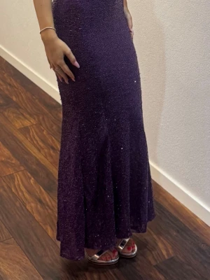Lång lila glittrig klänning - Säljer en elegant långklänning i lila med glittriga detaljer. Klänningen har en figurnära passform och är ärmlös, perfekt för bal eller fest. Materialet ger en lyxig känsla och den skimrar snyggt i ljuset. Perfekt för dig som vill sticka ut!