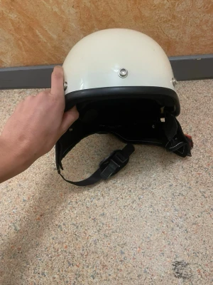 Vit retro hjälm med svart kant - Säljer en vit retroinspirerad hjälm med svart kant och detaljer i metall. Hjälmen har justerbart hakband och insida i svart vadderat material med vita sömmar. Perfekt för dig som vill ha en cool och klassisk look till moppe eller hoj.