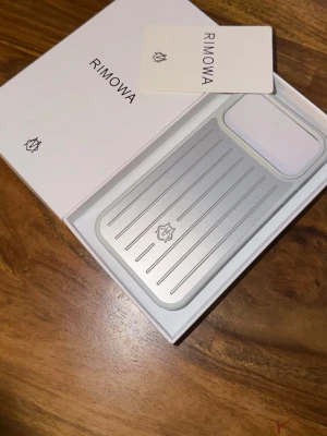 Iphone 17 pro case Rimowa - Snyggt mobilskal från RIMOWA i silverfärgad aluminium med räfflad design och vit detalj runt kameran. Skalets stilrena och moderna look gör det till ett riktigt statement för din mobil. Perfekt för dig som vill ha både skydd och design.