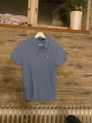 Blå pikétröja från Polo Ralph Lauren - Klassisk blå pikétröja från Polo Ralph Lauren med korta ärmar och den ikoniska broderade loggan på bröstet. Skjortan har krage och knappar vid halsen. Perfekt för en avslappnad men ändå stilren look.