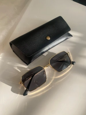 Solglasögon  - Helt nya Stilrena solglasögon med rektangulära svarta glas och tunna guldfärgade metallbågar. Kommer med svart fodral och putsduk. Perfekta för dig som gillar en clean och modern look.