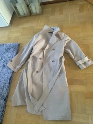 Beige trenchcoat med bälte - Stilren beige trenchcoat med dubbelknäppning och klassisk krage. Jackan har ett matchande bälte i midjan och dekorativa knappar vid ärmsluten. Tillverkad i ett lätt tyg som passar perfekt för lager-på-lager. Lång modell med slits bak för extra rörelsefrihet.