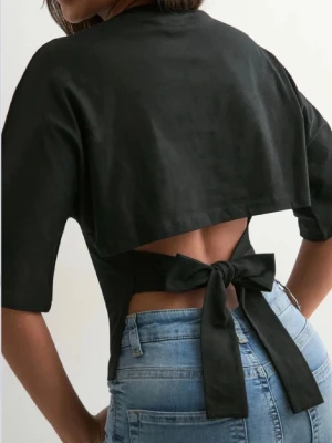 Svart croppad topp med knyt i ryggen - Trendig svart croppad topp med öppen rygg och breda knytband som ger en snygg rosett. Köpt från Nelly och är i bra skick, endast använd fåtal gånger.