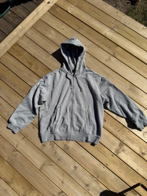 Ljusgrå zip hoodie med huva - En enkel och clean ljusgrå hoodie med dragkedja framtill och två fickor. Hoodien har justerbar huva med snören och ribbade muddar vid ärmar och nederkant. Perfekt för en avslappnad och chill stil. Materialet känns mjukt och bekvämt, troligen bomullsblandning.