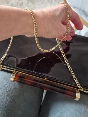 Svart lack kuvertväska med guldkedja - Elegant svart kuvertväska i glansigt lackmaterial med guldfärgad kedja och ett stilrent handtag i sköldpaddsmönster. Med innerficka. Väskan har en klassisk rektangulär form och passar perfekt till en trendig look. Kan bäras i handen ( kedja i vöskan) eller på axeln. Märket Dune.