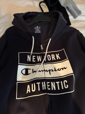 Svart hoodie från Champion med tryck - Säljer en svart hoodie från Champion med vit text på bröstet där det står 'New York Champion Authentic'. Hoodien har dragkedja, huva med vita snören och är långärmad. Perfekt för dig som gillar streetwear och vill ha en klassisk look.