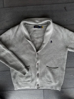 Ljusgrå kofta från Polo Ralph Lauren - Ljusgrå ribbstickad kofta från Polo Ralph Lauren med klassisk krage, knäppning framtill och två fickor. Ikoniska lilla loggan broderad på bröstet. Perfekt för lager på lager och enkel att matcha med olika outfits.