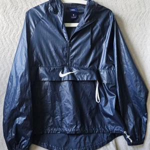 Mörkblå vindjacka från Nike - Snygg mörkblå vindjacka från Nike med halv dragkedja, huva och stor vit logga på bröstet. Jackan har en praktisk magficka och justerbar nederkant med dragsko. Tillverkad i lätt, glansigt material som står emot vind.