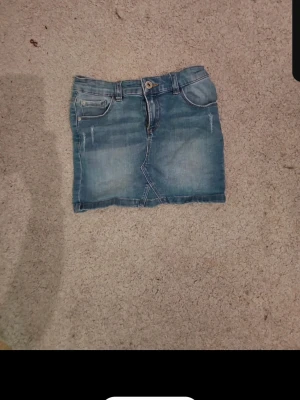 Blå jeanskjol  - Säljer en klassisk blå jeanskjol  med fickor både fram och bak samt knapp och dragkedja i midjan. Kjolen har en kort passform och är tillverkad i denim med snygga sömdetaljer. Perfekt för dig som gillar en avslappnad och trendig look.