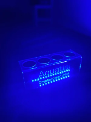 Aquatini LED-display med flaskhållare - Säljer en transparent rektangulär LED-display från Aquatini med texten 'Crystal clear drink'. Displayen har plats för fyra flaskor och lyser starkt blått. Materialet är plexiglas och designen är modern och stilren.