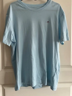 Ljusblå t-shirt från Gant - Säljer en ljusblå t-shirt från Gant med liten broderad logga på bröstet. Klassisk rund halsringning och korta ärmar. Tillverkad i mjuk bomull som känns skön mot huden. Perfekt till jeans eller shorts för en enkel och clean look.