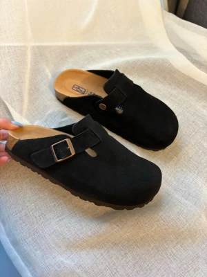 Svarta Birkenstock tofflor svart - Säljer ett par svarta Birkenstock tofflor i mjuk mocka med justerbart spänne i metall. Klassisk slip-in modell med bekväm fotbädd och räfflad sula. Perfekta för dig som vill ha både stil och komfort i vardagen. Vi kan diskutera priset