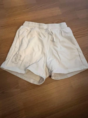 Vita shorts med snörning och strassdetalj - Säljer ett par vita shorts med snörning i midjan och en glittrig strassdetalj på ena benet. Mjukt material och avslappnad passform, perfekta för sommardagar eller chill hemma.